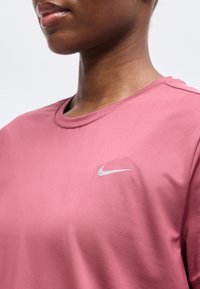 Γυναίκα που φοράει ροζ μπλούζα Nike με μικρό λευκό λογότυπο Swoosh στο στήθος, εμφανίζεται από το λαιμό έως τον άνω κορμό.