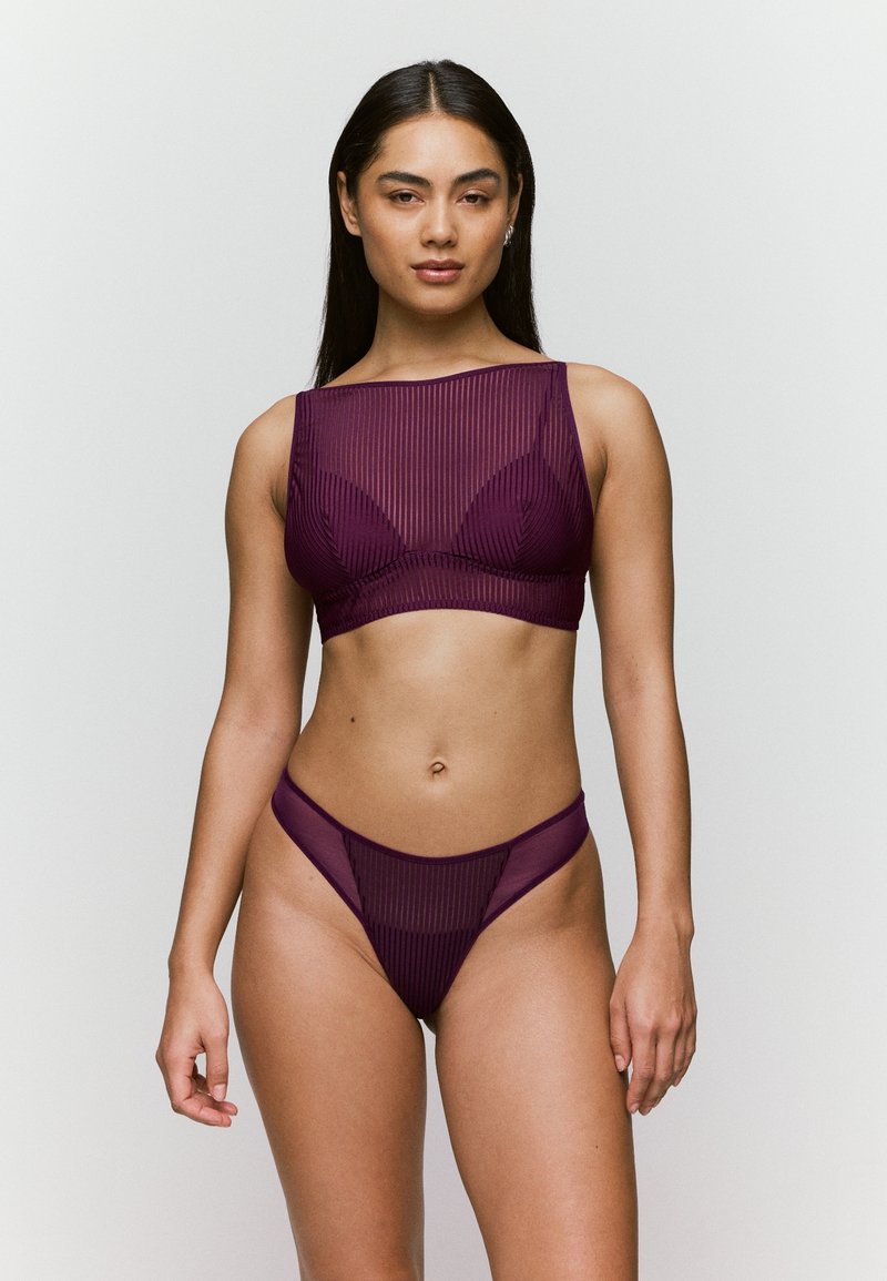 Vrouw in een doorschijnende pruimenkleurige gestreepte lingerie-set met een crop top en hoog uitgesneden ondergoed, staand tegen een effen lichte achtergrond.