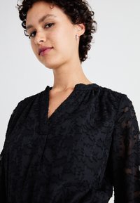 Juoda bluzelė su tekstūruota gėlių siuvinėjimu, subtiliu V formos iškirpimu ir permatomomis ilgomis rankovėmis. Lygus audinys su sudėtingu apdailos motyvu.