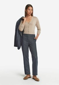 Beige knapplöst cardigan över mörkblå byxor med en subtil struktur. Modellen står med handen i fickan och har på sig bruna sandaler.