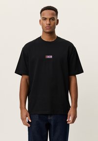 Νέος άνδρας με μαύρο φαρδύ T-shirt με μικρό ροζ λογότυπο "LES DEUX" στο στήθος και σκούρο μπλε τζιν, που στέκεται μπροστά σε απλό φόντο.