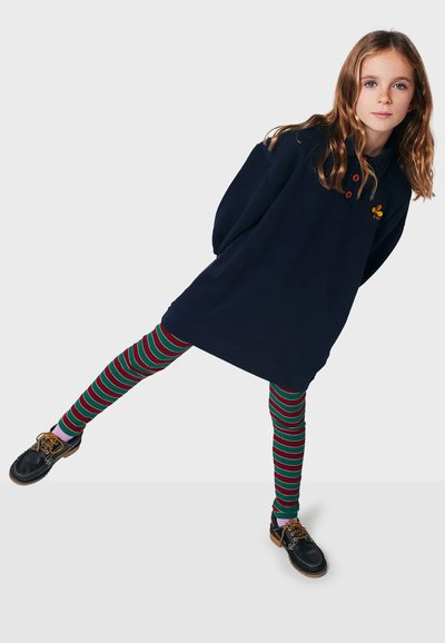 Robe en polaire marine avec des accents de boutons rouges et un logo brodé, associée à des leggings rayés verts et rouges et des chaussures sombres.