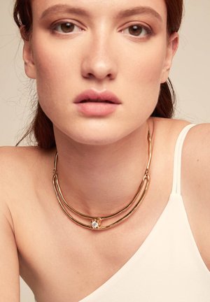 Collar de metal en tono dorado con dos hebras curvas, que presenta un adorno central con una piedra transparente. Textura suave y pulida.