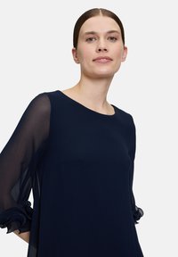 Robe bleu marine en tissu lisse, avec des manches transparentes et des accents volantés aux poignets. Encolure ronde simple et classique.