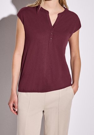 Kvinde iført en kortærmet burgunder henley-top med tre knapper og beige bukser, hænder afslappet ved siderne.