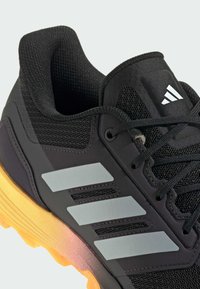 Černá sportovní bota s síťovaným svrškem, stříbrnými pruhy a oranžovou podrážkou. Má zaoblený tvar, texturovaný povrch a zesílenou patu.