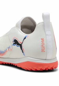 Biele športové topánky s textúrovaným vrchom, čiernym logom Puma, oranžovou podrážkou a červenými/modrými akcentmi. Design na obúvanie, hladký a odolný materiál.