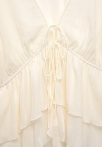 Détail d'une robe couleur crème avec décolleté en V, tissu froncé, volants superposés et cordons noués au centre devant.