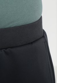 Svarta byxor med ett brett, texturerat midjeband. Tyget verkar slätt och något stretchigt. Toppen är i en dämpad grön färg som visar sömmen.