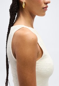 Γυναίκα με μακριά πλεξούδα, που φοράει υφασμένο λευκό αμάνικο τοπ και μεγάλο χρυσό σκουλαρίκι σε σχήμα δάκρυ, εμφανιζόμενη σε προφίλ.