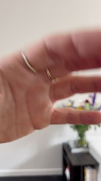 Primer plano de una mano con dos anillos de oro delgados. Se puede ver una textura suave de la piel. Al fondo, una disposición borrosa de flores coloridas.