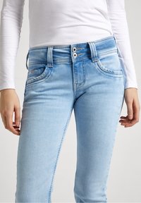 Ljusblå denimjeans med en figursydd design, stängning med två knappar vid midjan, framfickor och subtila sömnadsdetaljer längs sömmarna.