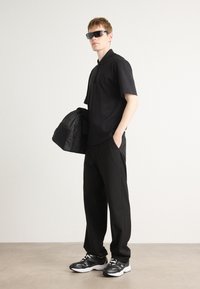 Svart kortermet polo skjorte med glidelås i kragen, svarte vide bukser og svart-hvite joggesko, accessorisert med mørke solbriller.