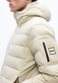 Άνδρας φοράει μπεζ επενδεδυμένο μπουφάν με κουκούλα, που διαθέτει τσεπάκι με φερμουάρ και μαύρο λογότυπο "BOSS" στο επάνω αριστερό μανίκι.