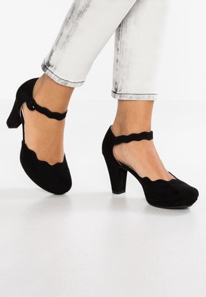 Sorte ruskind Mary Jane-hæle med bølgede kanter, båret med opkørte, lysevaske jeans på en hvid reflekterende gulv.