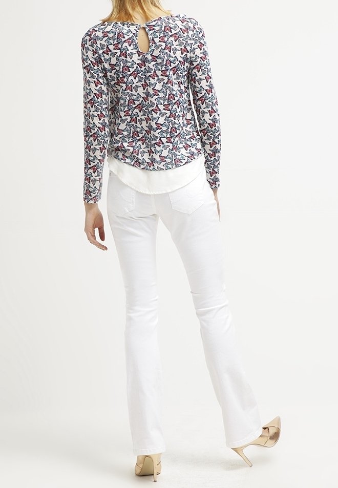 Femme debout, portant un haut à manches longues avec un imprimé papillon et une ouverture en forme de clé dans le dos, une blouse blanche superposée, un jean blanc et des talons beiges.