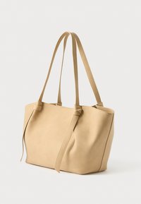 Beige læder tote taske med to lange håndtag, en blød, struktureret form og knyttede detaljer på siderne. Glat tekstur med minimale designdetaljer.