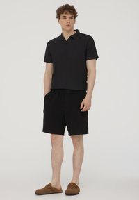 Polo nero a maniche corte con scollo a V, abbinato a pantaloni corti neri. Scarpe marroni senza lacci completano l'outfit.