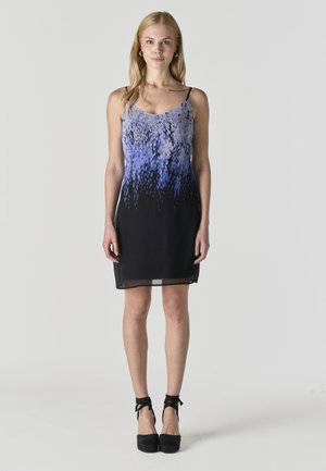 Mladá žena stojaca, oblečená v šatách bez rukávov s abstraktným modro-čiernym vzorom a čiernymi platformovými topánkami viazanými okolo členkov.