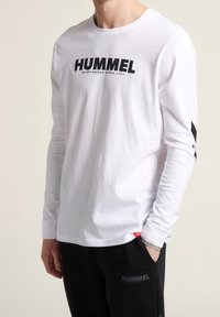 Witte blouse met lange mouwen, featuring een groot zwart "HUMMEL" logo op de voorkant en diagonale zwarte strepen op de mouwen. Katoenen stof, casual pasvorm.