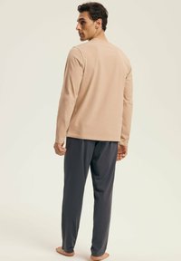 Man standing barefoot, facing away, wearing a beige long-sleeve shirt and dark gray loose pants against a plain light background.