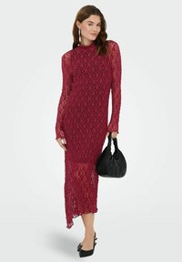 Robe en dentelle bordeaux à manches longues et col haut, avec un corsage ajusté et un ourlet qui descend en biais. Tient un sac à main noir.