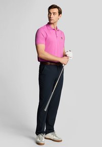 Pink polo shirt with a logo on the chest, navy trousers, and white shoes. The model holds a golf club with a gloved hand.