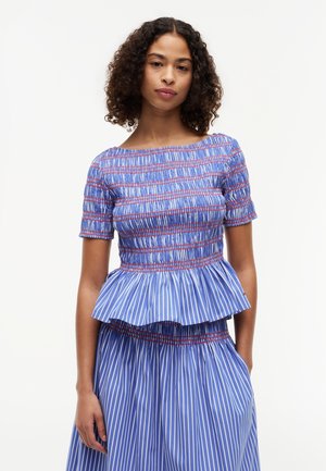 Junge Frau mit lockigem Haar, die ein blau-weiß gestreiftes Peplum-Top mit roter Naht und passenden Rock trägt, vor weißem Hintergrund stehend.
