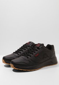 Schwarze Sportschuhe aus synthetischem Leder mit Schnürdesign, roten Logoakzenten, durchbrochener Verarbeitung und Gummisohle.