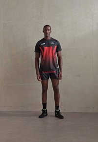 Mann in schwarzer und roter Sportbekleidung, der vor einer schlichten grauen Wand steht und schwarze High-Top-Sneaker sowie schwarze Socken trägt.