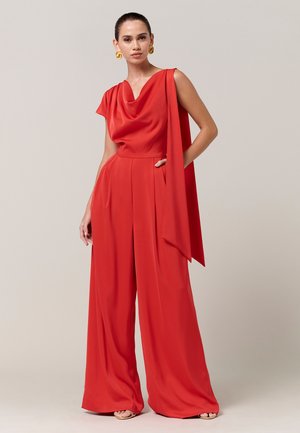 Žena v červenom bezrukávovom overal s voľnými nohavicami a záhybovým výstrihom, so zlatými špirálovými náušnicami, stojaca na jednoduchom svetlom pozadí.