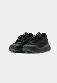 Paire de baskets noires avec semelles épaisses texturées, lacets et logo discret de la marque sur le côté et la languette, sur fond blanc.