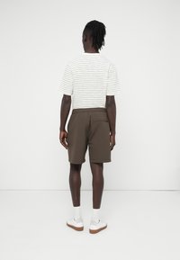 Καφέ σορτς με ελαστική μέση και πίσω τσέπη, συνδυασμένα με λευκό πουκάμισο με ρίγες. Το μοντέλο φοράει λευκά παπούτσια και κάλτσες.