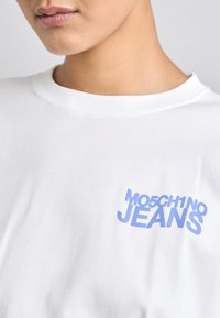 Balta kokvilnas T-kreklu ar zilu grafisku izdrukā, kas uzraksta "MO5CH1NO JEANS" kreisajā krūšu pusē. Īsas piedurknes un apaļš kakla izgriezums.