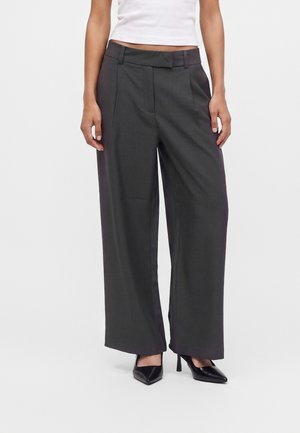 Femme portant un pantalon gris foncé taille haute, jambes larges, avec passants de ceinture, associé à un top court blanc et des chaussures à talons noirs.