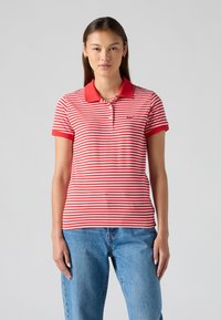 Polo shirt rayé rouge et blanc avec un col et des manches courtes, comportant un petit logo sur la poitrine gauche. Porté avec un jean bleu.