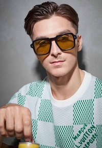 Mustat suorakulmaiset aurinkolasit keltaisilla linsseillä, käytettynä valkoisen ja vihreän kuvioidun paidan kanssa, jossa on teksturoitu shakkilautakuvio.