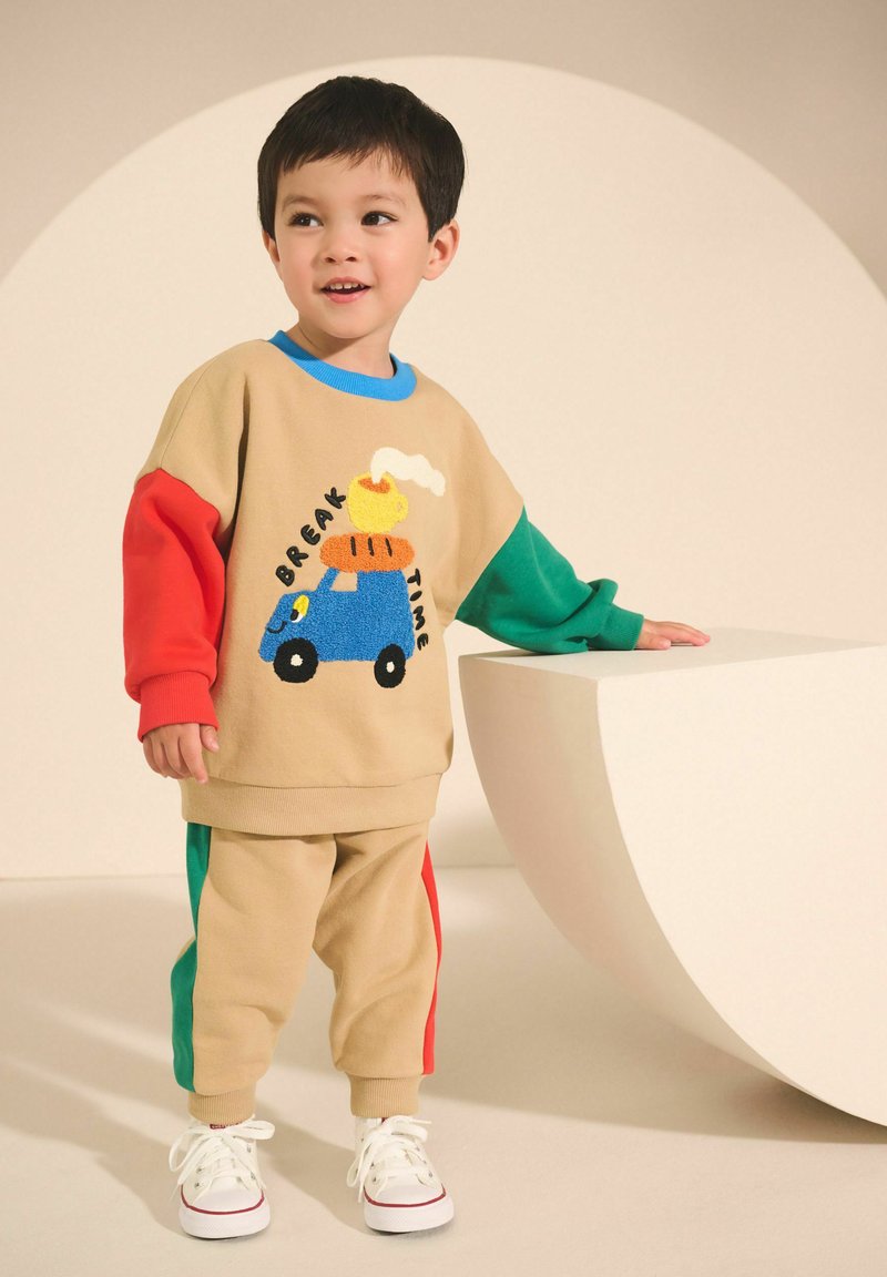 Jeune garçon debout et souriant, portant une tenue beige avec des manches rouges et vertes, et un motif de voiture bleue avec texte sur le sweat-shirt.