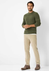 Olivgrüner Strickpullover mit gerippten Bündchen und Saum, kombiniert mit hellbeigen Cordhosen und braunen Lederstiefeln.