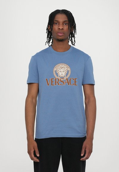 Tricou din bumbac albastru, cu guler rotund și mâneci scurte. Pe partea din față, în centrul tricoului, se află un logo imprimat cu Medusa, iar sub acesta este textul "VERSACE".