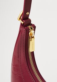 Burgundy leather bag with a shiny finish, gold zipper, and branding tag. Features a curved shape and detailed stitching.