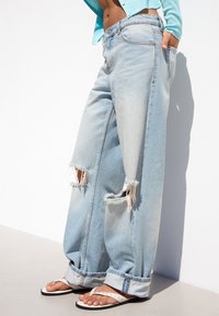 Jean bleu clair déchiré à jambes larges porté avec un cardigan court turquoise et des sandales blanches sur un mannequin sur fond blanc.