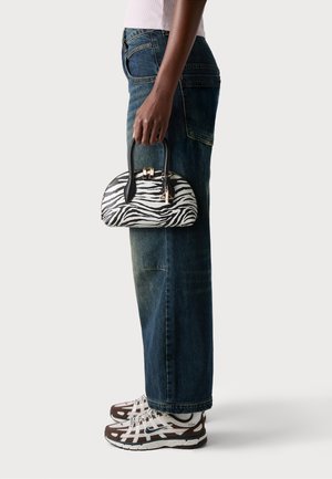 Person med vida jeans och sneakers, som håller en liten handväska med svart-vitt zebramönster och svarta handtag.
