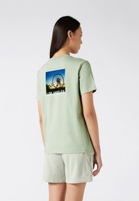 Camiseta de algodón verde con un gráfico cuadrado de una noria y el texto "LOS ÁNGELES" en la parte posterior. Corte relajado, mangas cortas y textura suave.