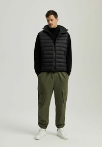 Czarna kamizelka puchowa z kapturem noszona na czarnym golfie, w połączeniu z oliwkowymi spodniami cargo z bocznymi kieszeniami i elastycznymi mankietami, uzupełniona białymi trampkami.