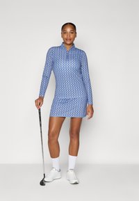 Blue patterned golf dress with long sleeves and collar, made from stretchy fabric. Model holds a golf club, wearing white shoes and socks.