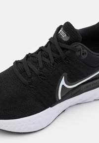 Zapatilla deportiva negra con una parte superior de punto texturizado, mediasuela blanca y un prominente logo de Nike en negro. Presenta cordones planos y un cuello acolchado.