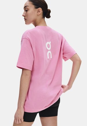 Jeune femme aux cheveux foncés attachés en chignon, portant un T-shirt rose ample avec un logo blanc dans le dos et un short noir, vue de profil.