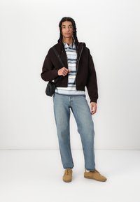 Haut à capuche noir court, chemise rayée bleue et blanche, jeans bleu clair et baskets beiges. Un petit sac bandoulière noir souligne l'ensemble.