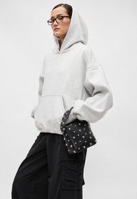 Siv hooded sweatshirt z sprednjo torbico, v kombinaciji z črno torbo s srebrnimi konicami, oblečeno z ohlapnimi črnimi hlačami.